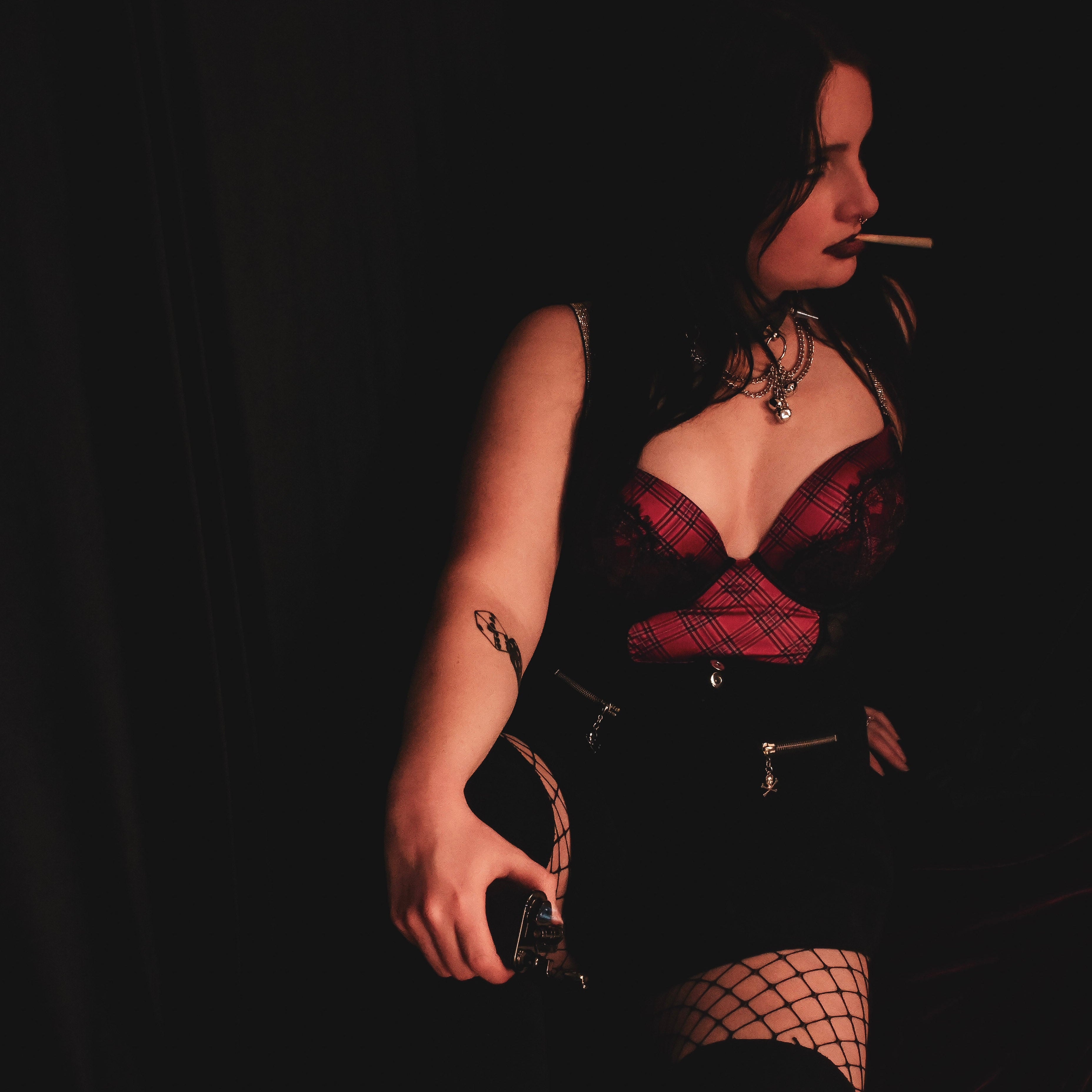 A woman in a dark gothic outfit featuring a red and black lace corset top, black skirt, fishnet stockings, and red lace-up boots, holding a cigarette against a very dark background.