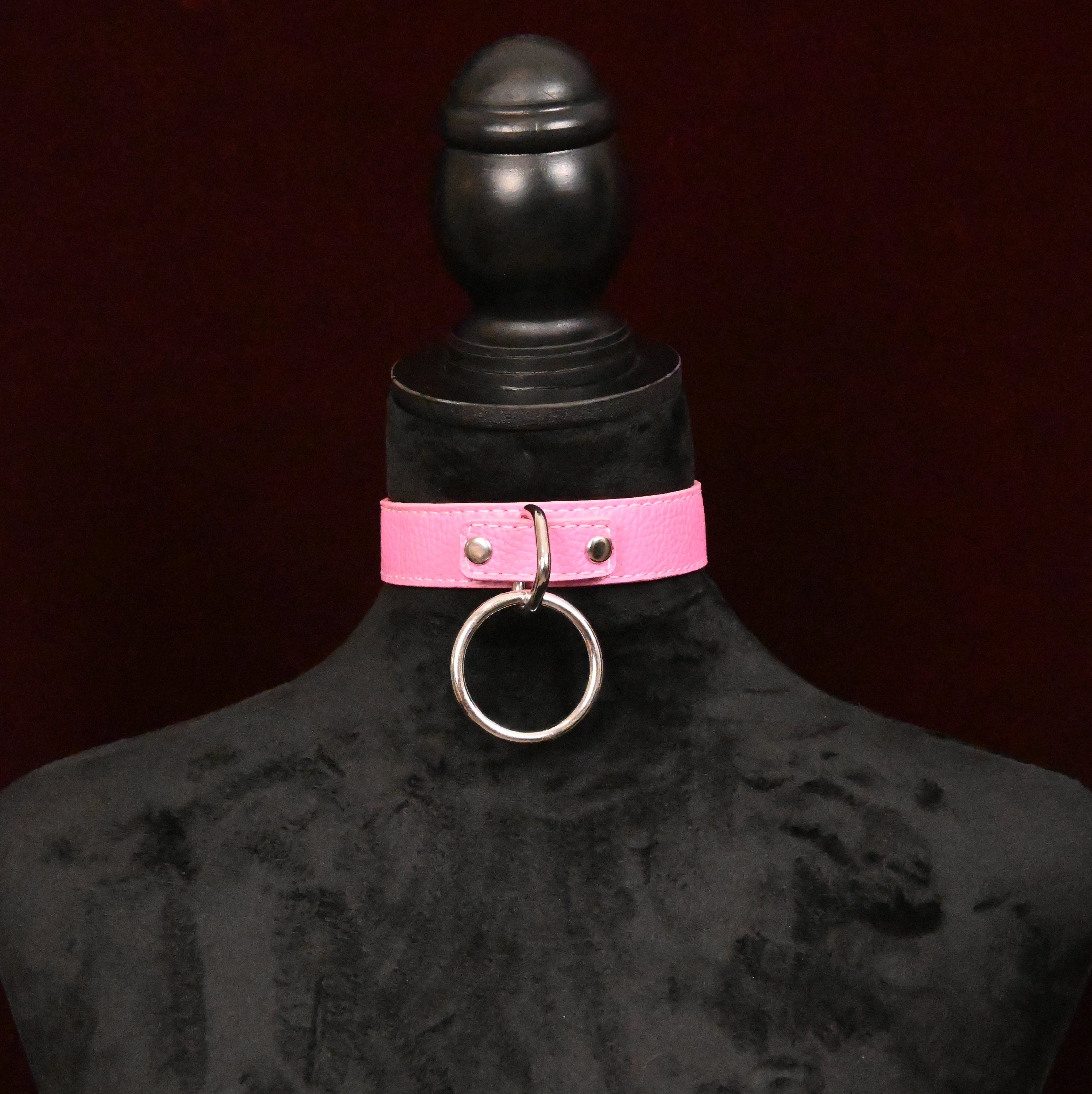 Pink leather collar with silver O ring on a black mannequin against a dark red background
