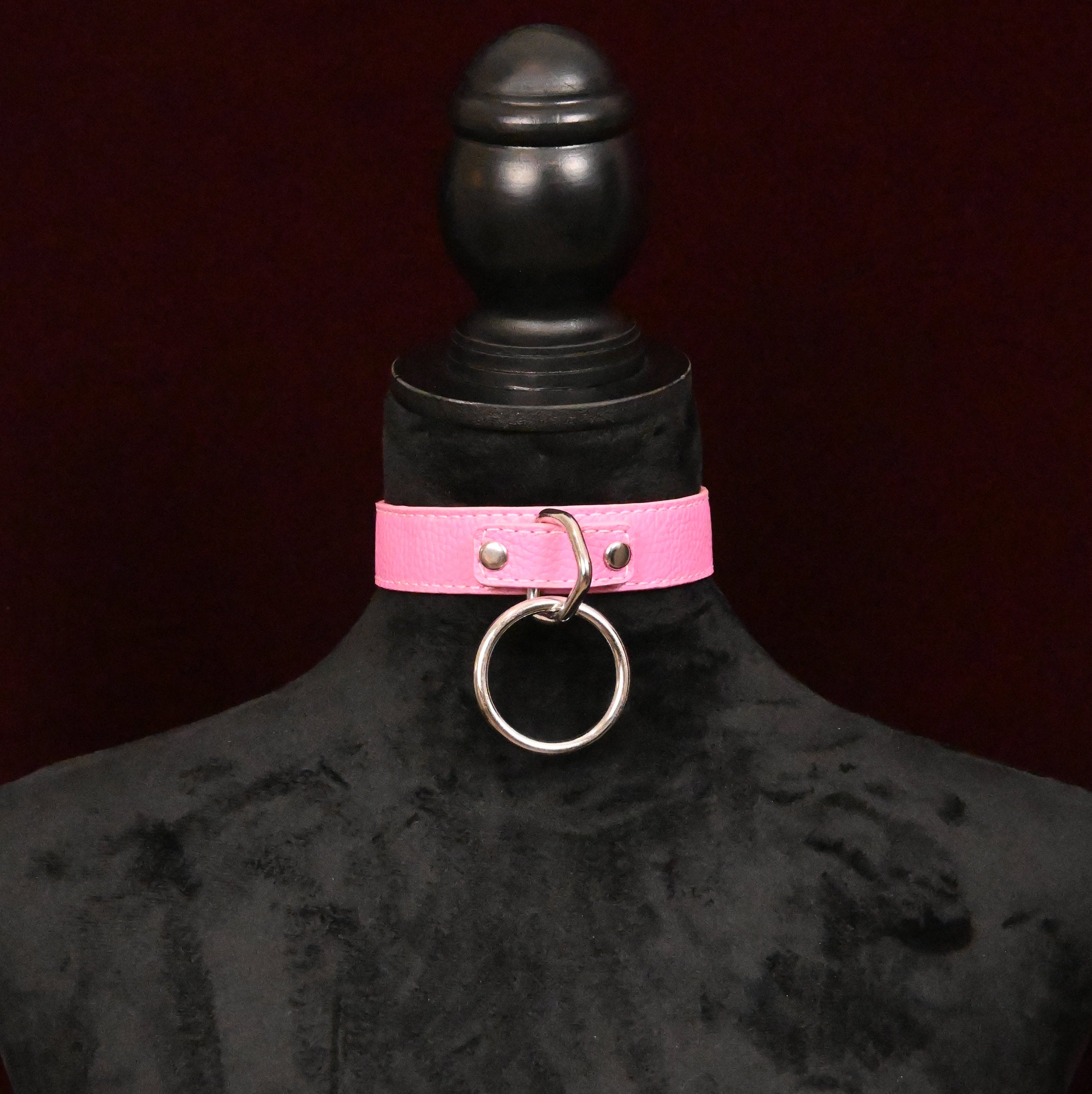 Pink leather choker with silver O ring on a black mannequin against a dark red background