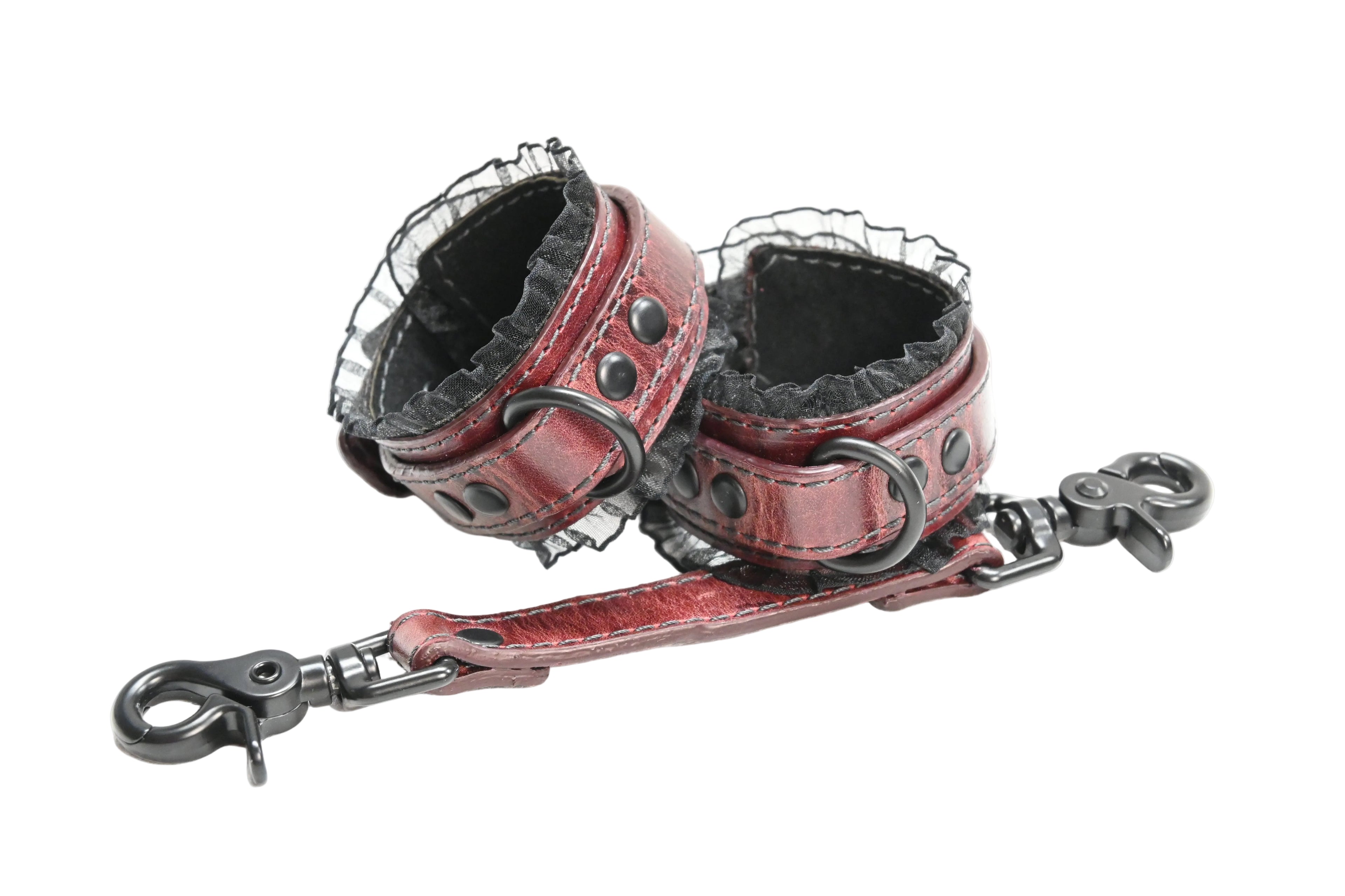 A pair of red leather wrist cuffs with black lace trim, black suede lining, and black metal rings and rivets, photographed on a plain white background. Cuffs are photographed with a matching red leather link with clasps on each end.