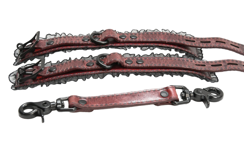 A pair of red leather wrist cuffs with black lace trim, black suede lining, and black metal rings and rivets, photographed on a plain white background. Cuffs are photographed with a matching red leather link with clasps on each end.