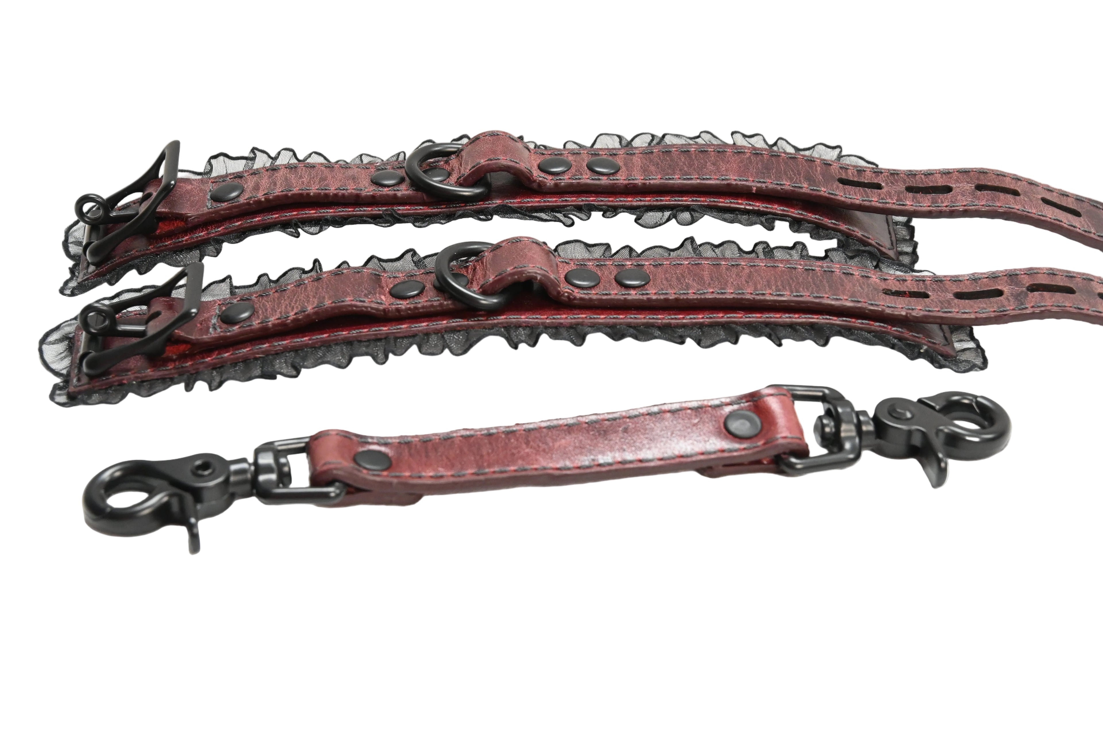 A pair of red leather wrist cuffs with black lace trim, black suede lining, and black metal rings and rivets, photographed on a plain white background. Cuffs are photographed with a matching red leather link with clasps on each end.