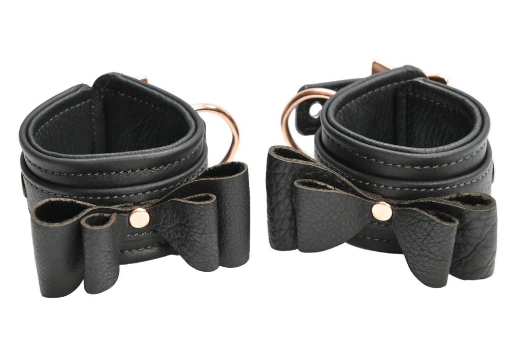 Two black leather wrist cuffs with bow details and rose gold metal hardware, displayed on a clean white background.
