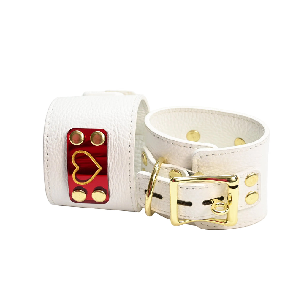 Two white leather cuffs with gold hearts on red heart acrylic plates, gold rivets, and gold D rings, photographed on a clean white background.