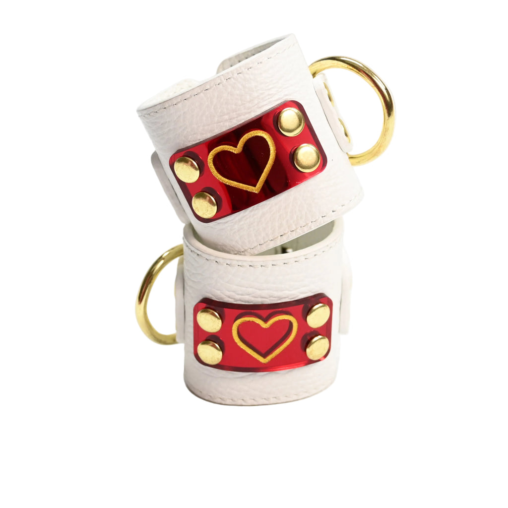 Two stacked white leather cuffs with gold hearts on red heart acrylic plates, gold rivets, and gold D rings, photographed on a clean white background.