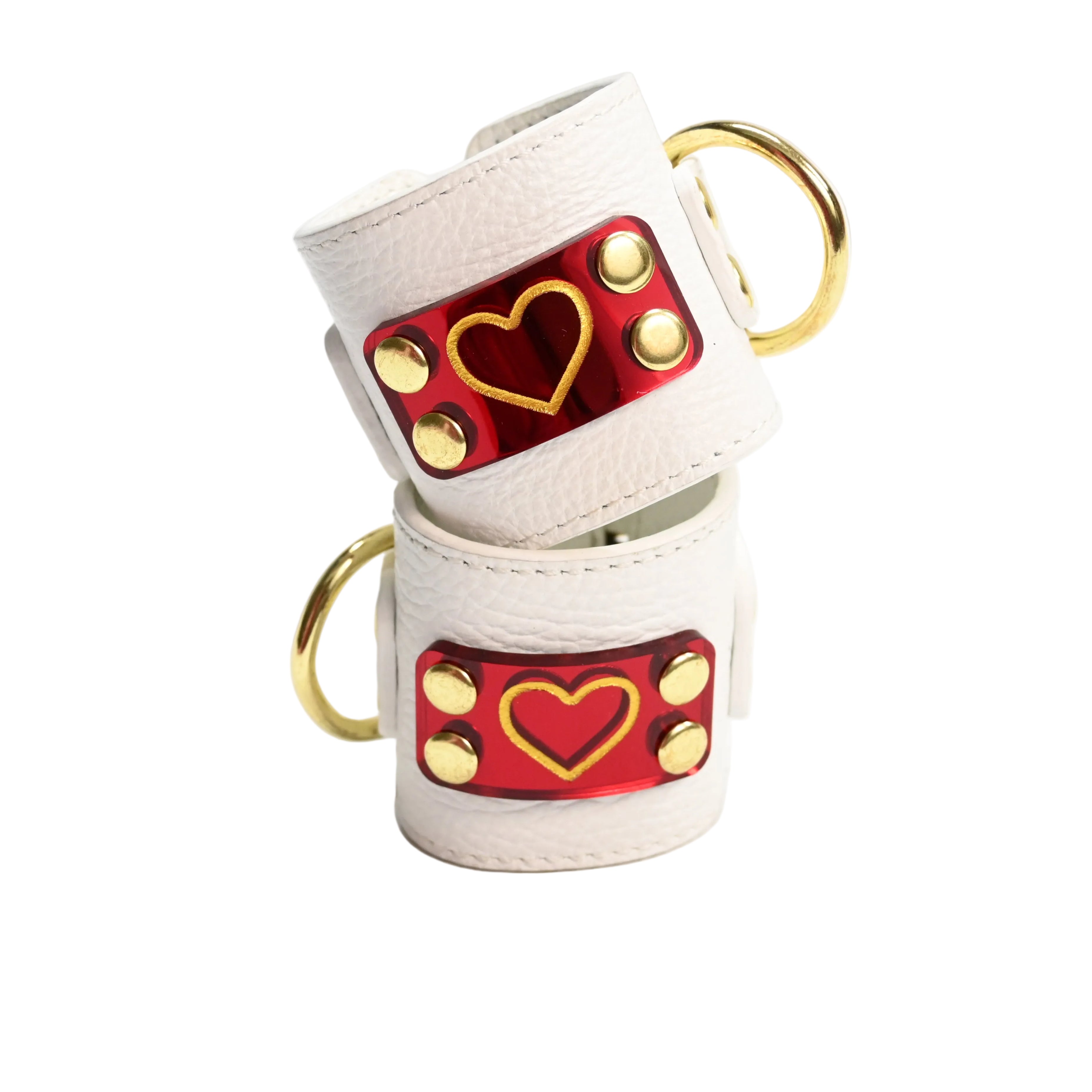 Two stacked white leather cuffs with gold hearts on red heart acrylic plates, gold rivets, and gold D rings, photographed on a clean white background.