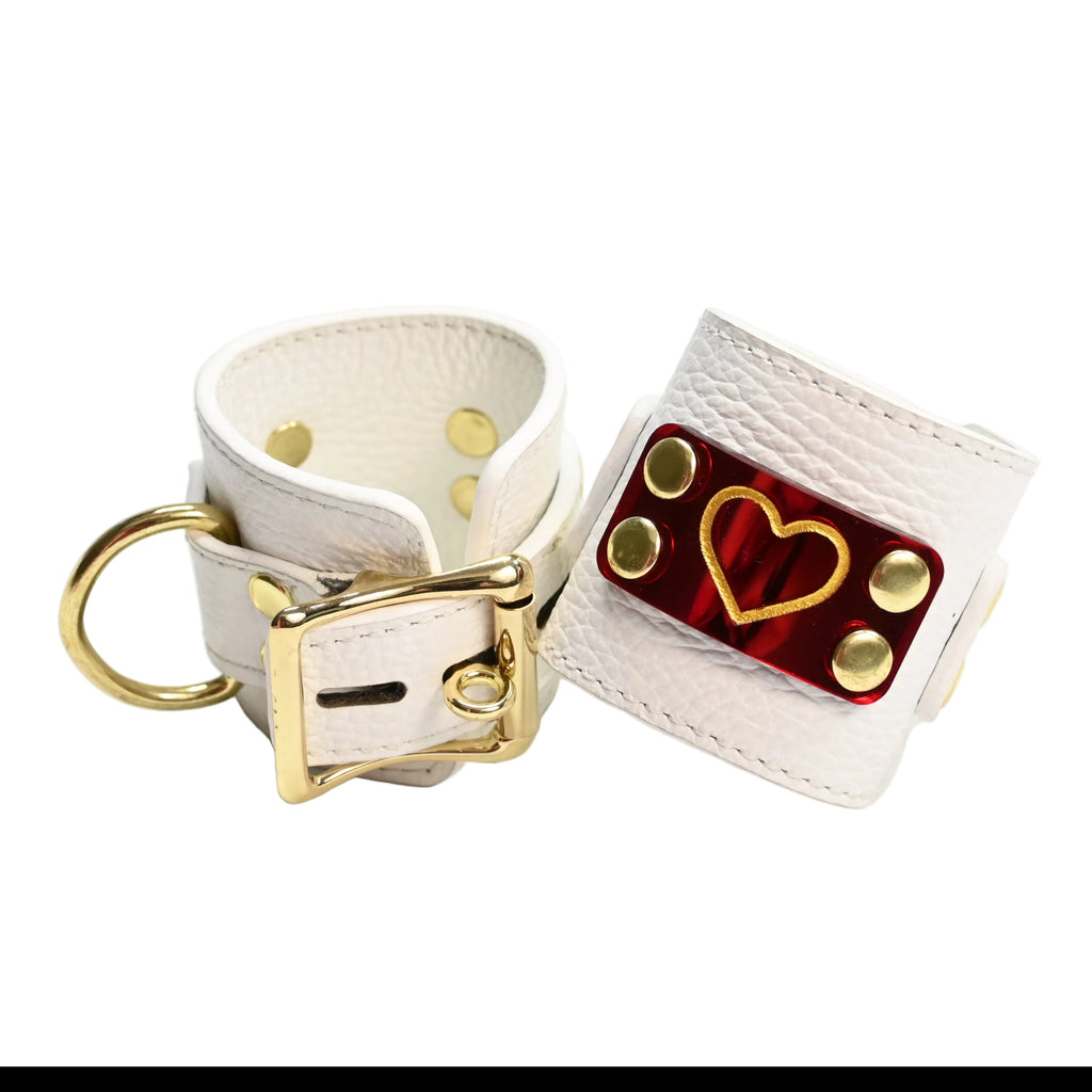 Two white leather cuffs with gold hearts on red heart acrylic plates, gold rivets, and gold D rings, photographed on a clean white background.
