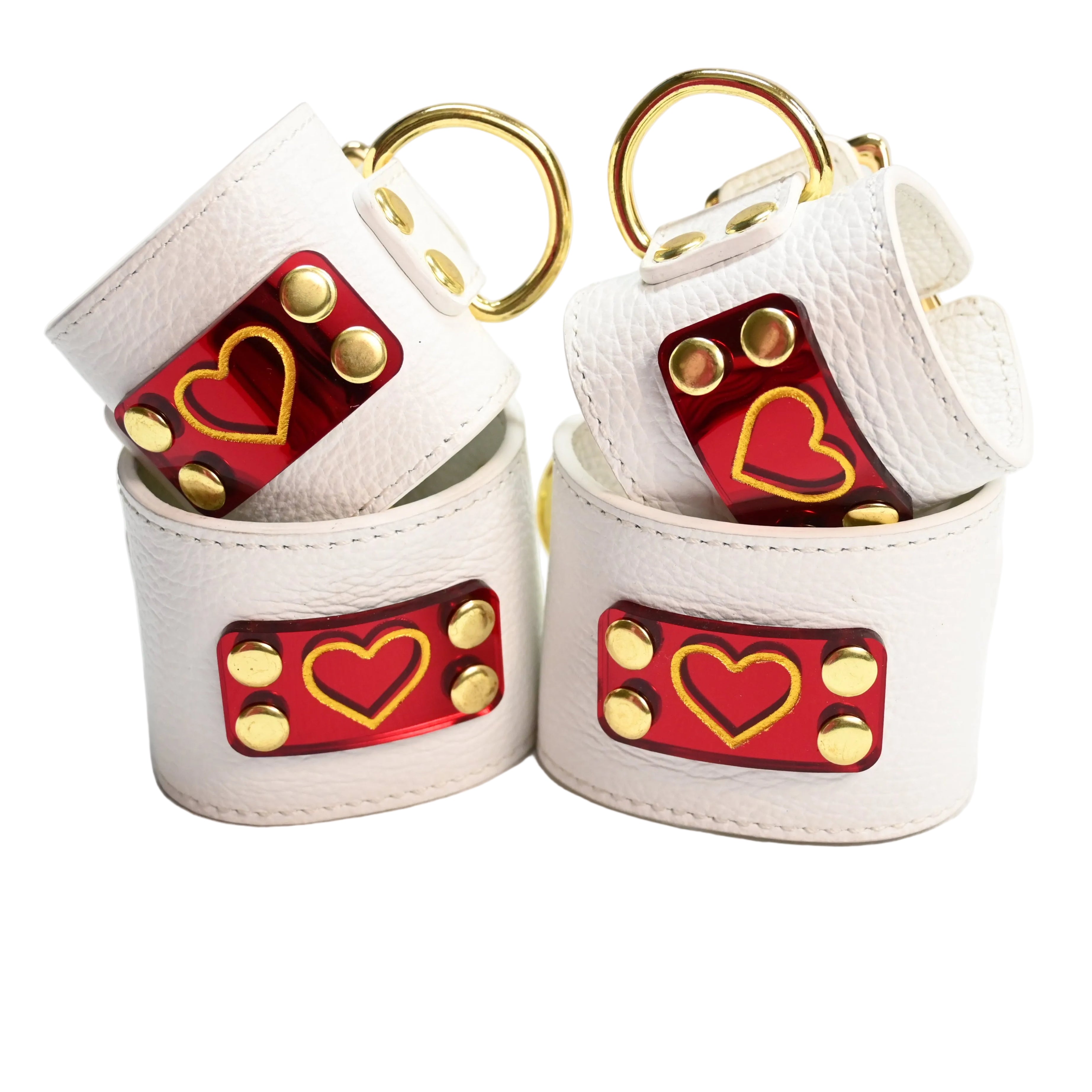 Two sets of white leather cuffs with gold hearts on red heart acrylic plates, gold rivets, and gold D rings, photographed on a clean white background.