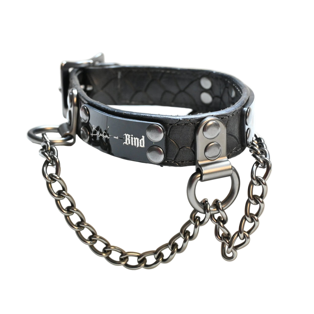 A black textured leather collar with silver hardware, featuring a chain and a plate engraved with 'Blush and Bind'  displayed on a white background.