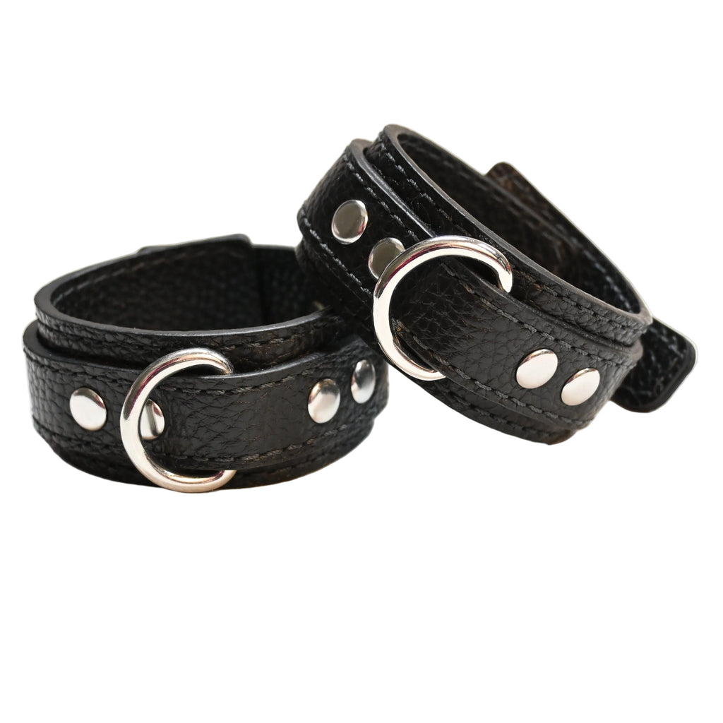 Two black leather cuffs with silver metal D rings and rivets, displayed on a clean white surface.