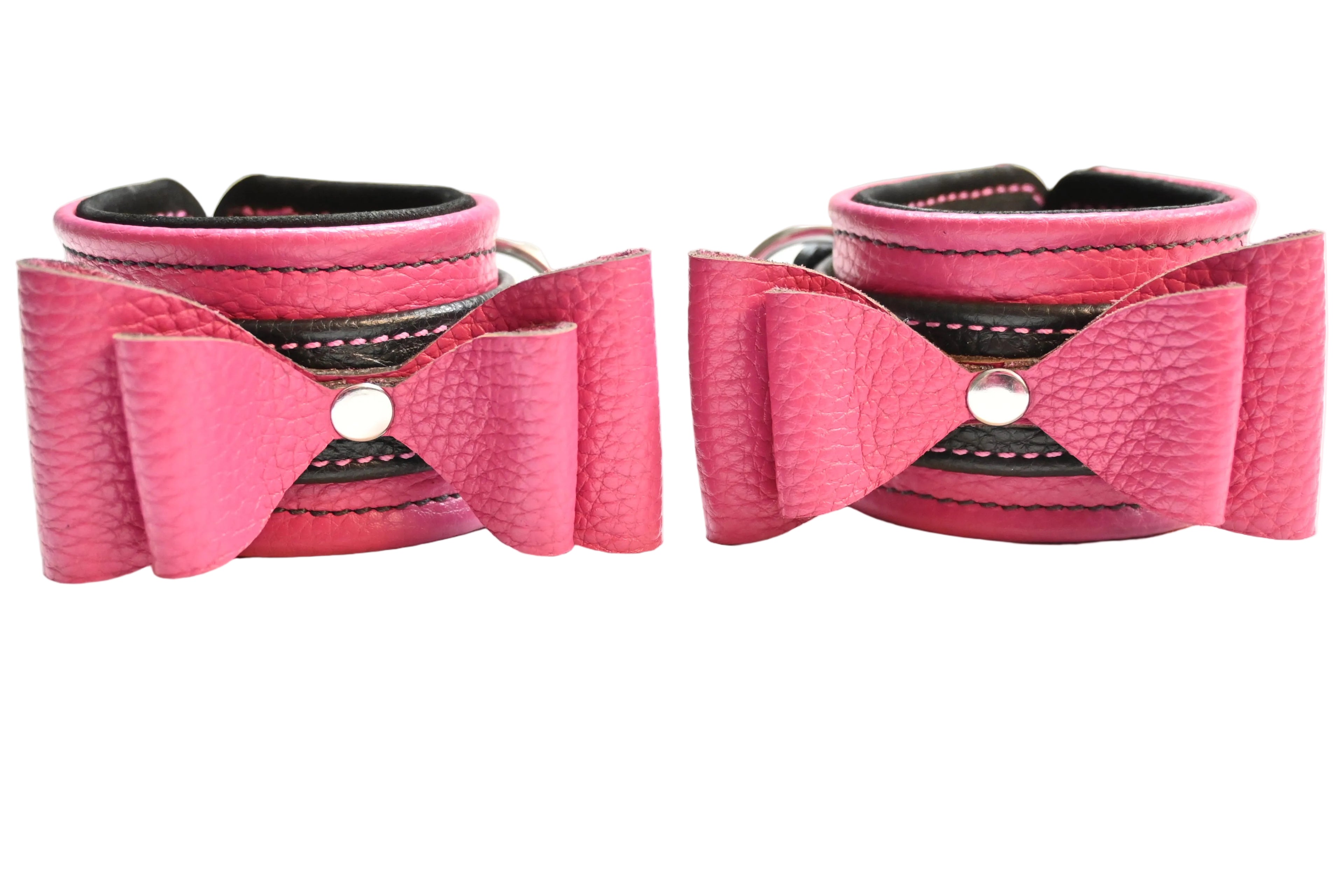 Two pink and black leather bow cuffs with black lining and silver hardware, displayed side-by-side on a white background.