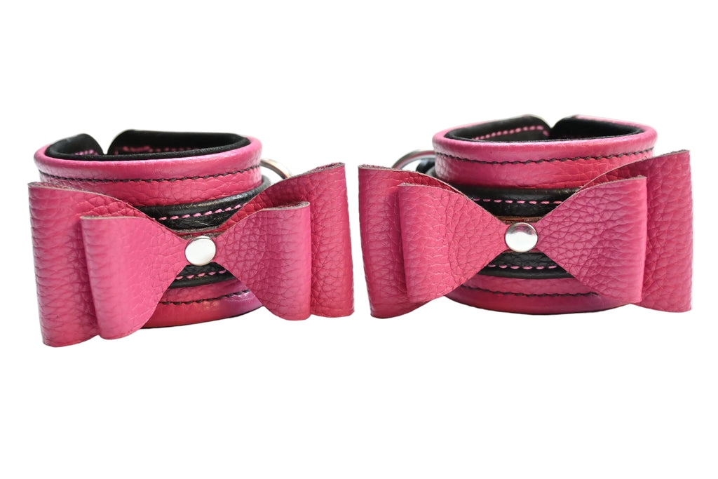 Two pink and black leather bow cuffs with black lining and silver hardware, displayed side-by-side on a white background.
