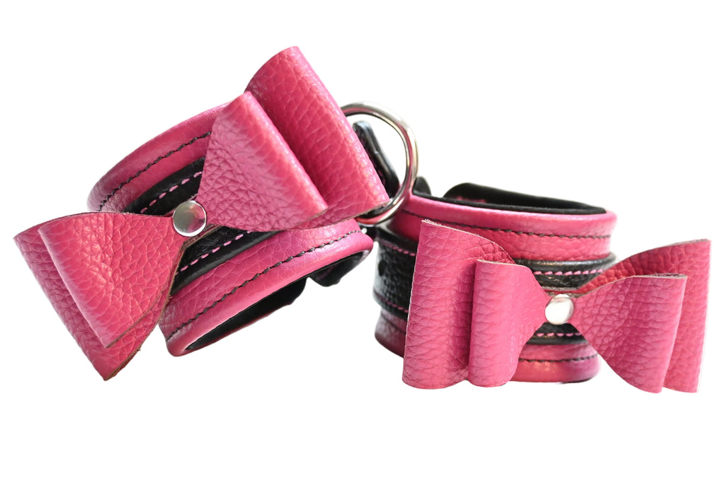 Two pink and black leather bow cuffs with black lining and silver hardware, displayed side-by-side on a white background.