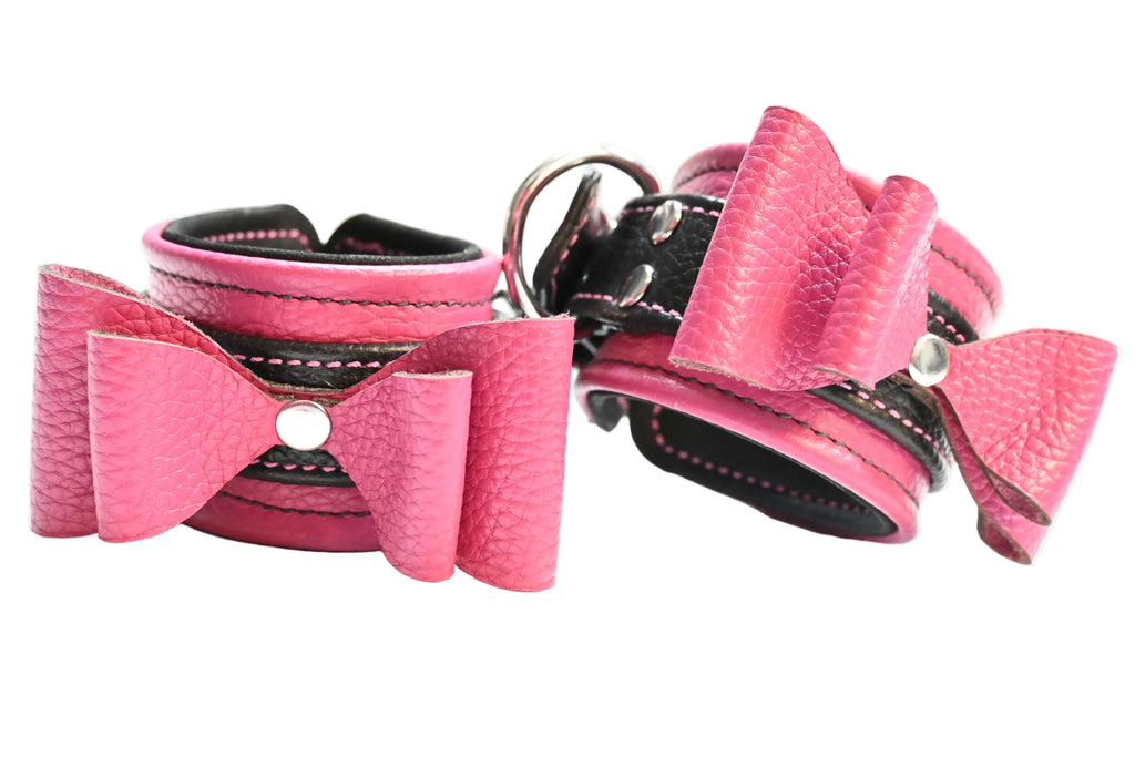 Two pink and black leather bow cuffs with black lining and silver hardware, displayed side-by-side on a white background.