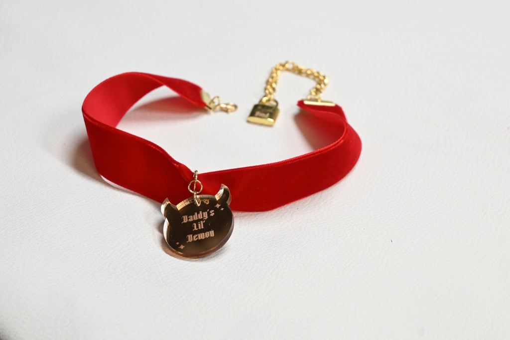 A red velvet choker collar with gold-tone hardware, featuring a pendant engraved with 'Daddy's lil' demon' and a gold chain clasp.