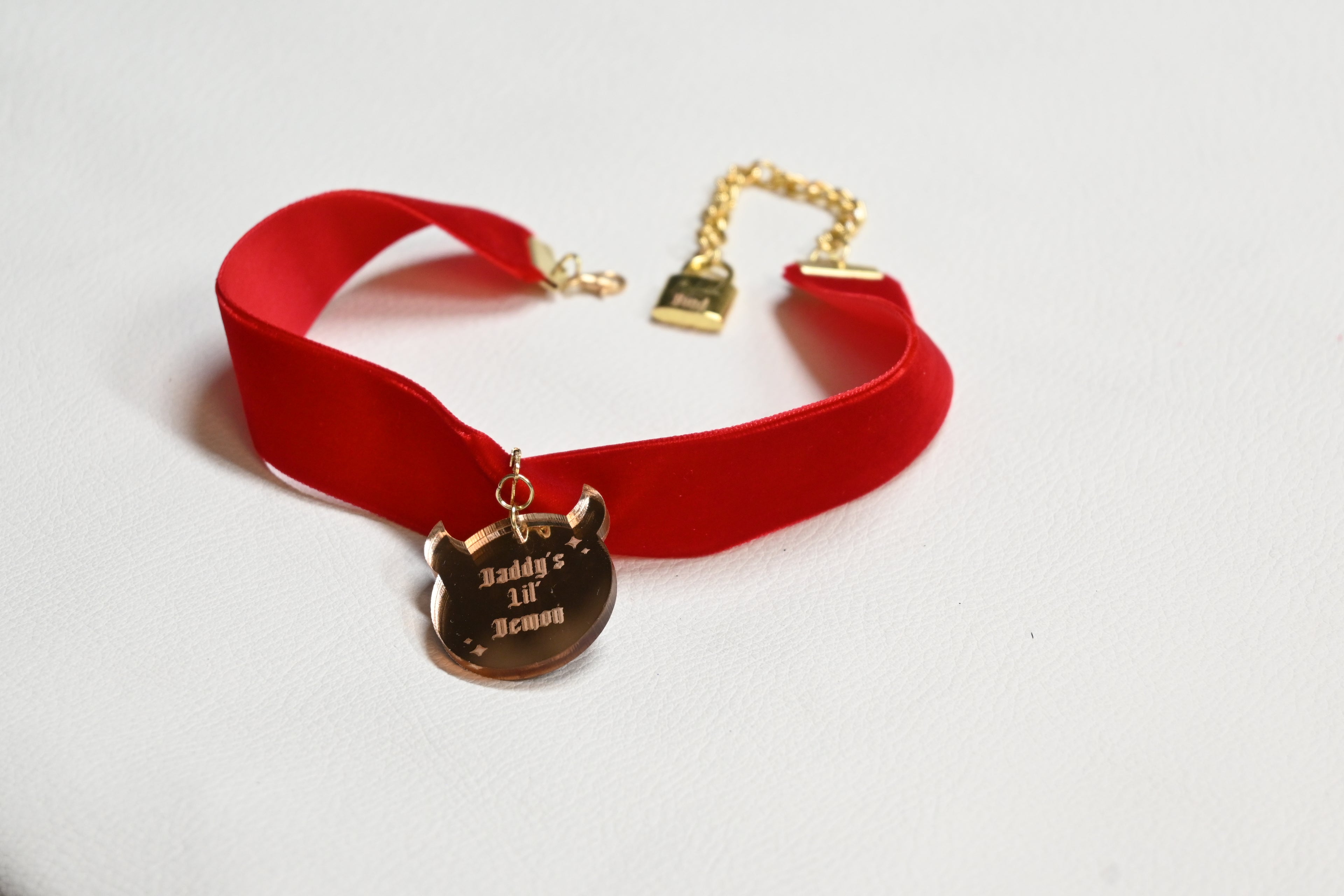 A red velvet choker collar with gold-tone hardware, featuring a pendant engraved with 'Daddy's lil' demon' and a gold chain clasp.