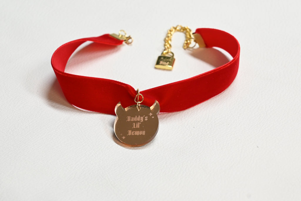 A red velvet choker collar with gold-tone hardware, featuring a pendant engraved with 'Daddy's lil' demon' and a gold chain clasp.