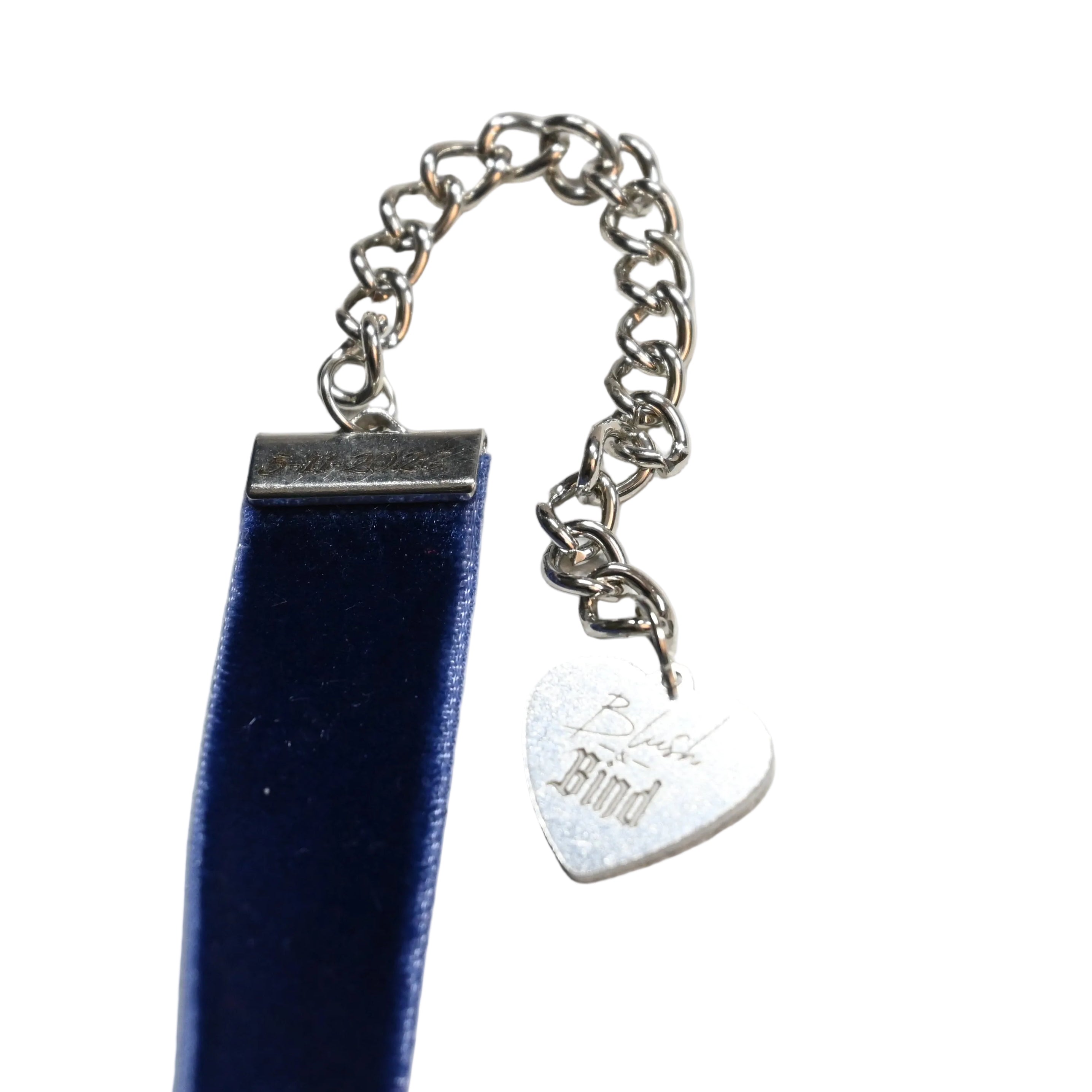 A close-up of a silver chain accessory with a heart-shaped engraved tag, shown with dark blue velvet choker on white background.
