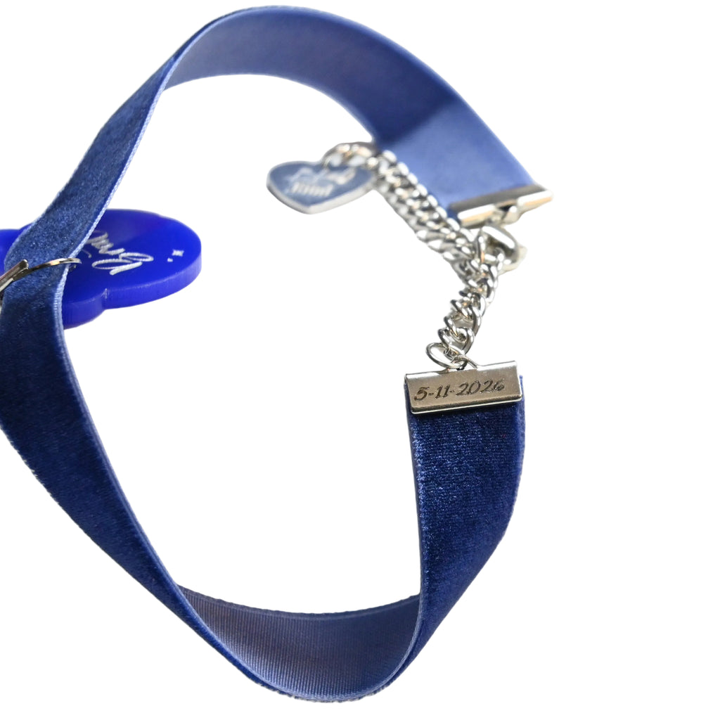 A close-up of a blue velvet ribbon choker with a silver chain extender and a small rectangular metal tag engraved with a date.