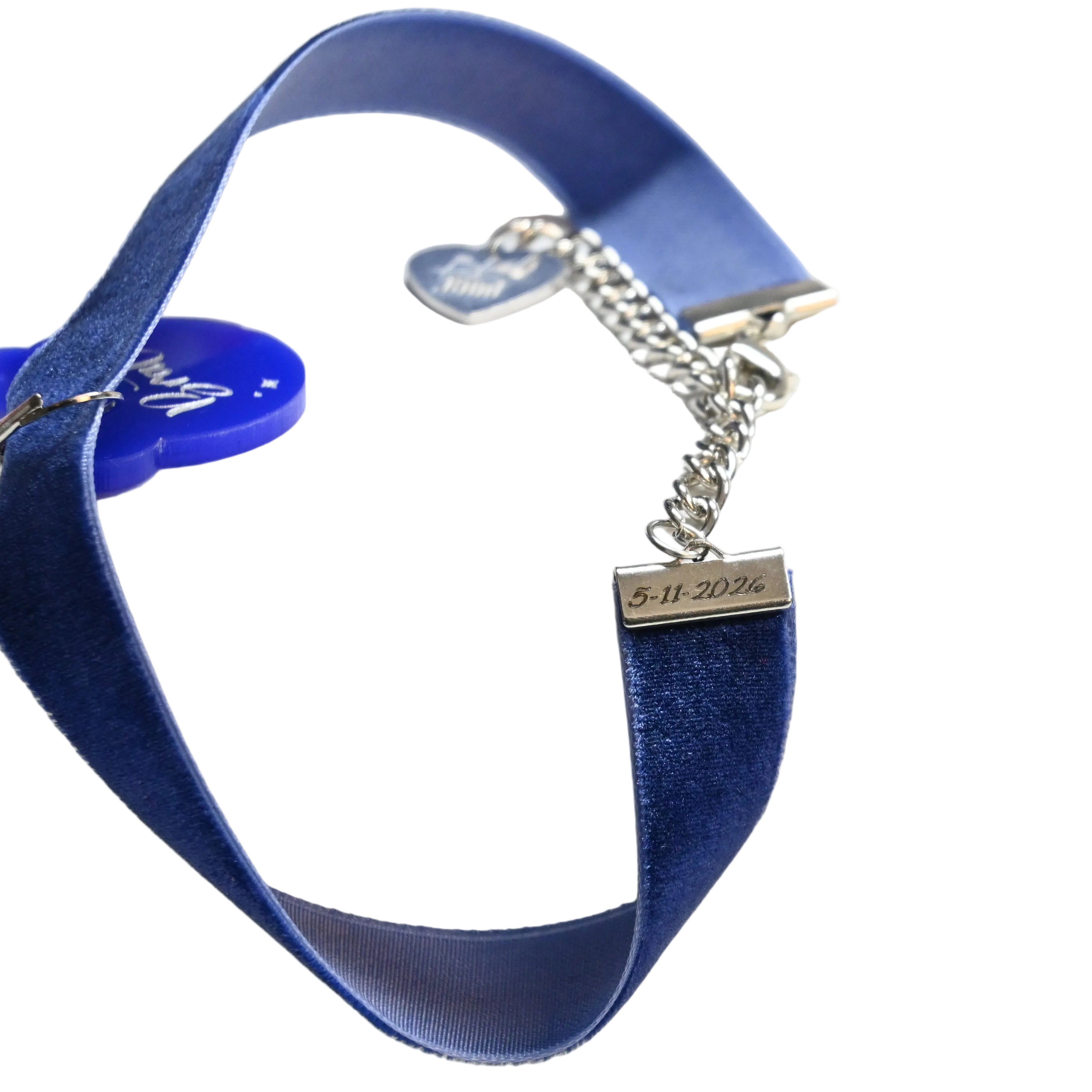 A close-up of a blue velvet ribbon choker with a silver chain extender and a small rectangular metal tag engraved with a date.