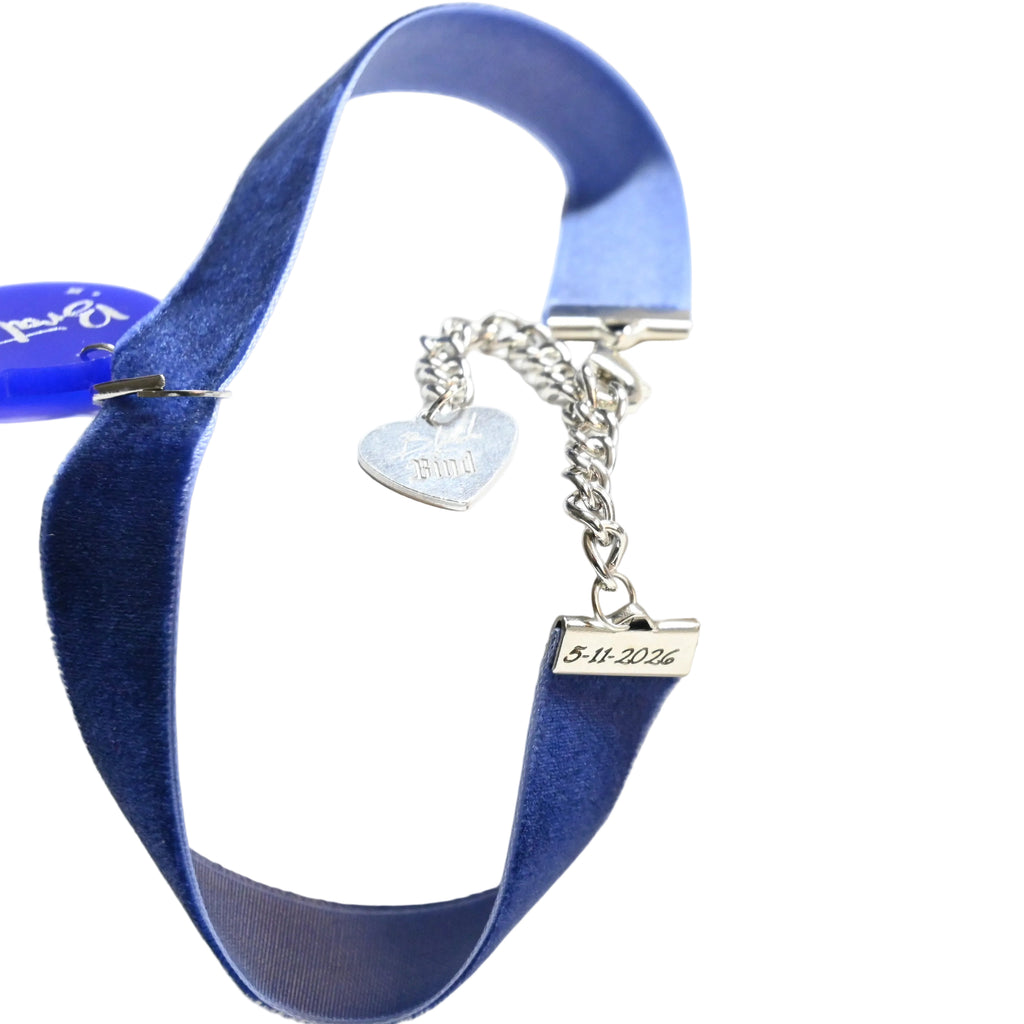 A blue velvet ribbon bracelet with a silver-tone chain featuring a heart-shaped charm and a small engraved tag, displayed on a white surface.