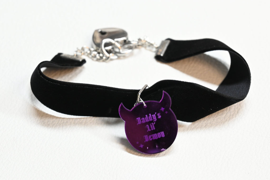 A black velvet collar with a silver clasp and a purple demon-shaped charm engraved with 'Daddy's LIL' Demon', displayed on a white background.